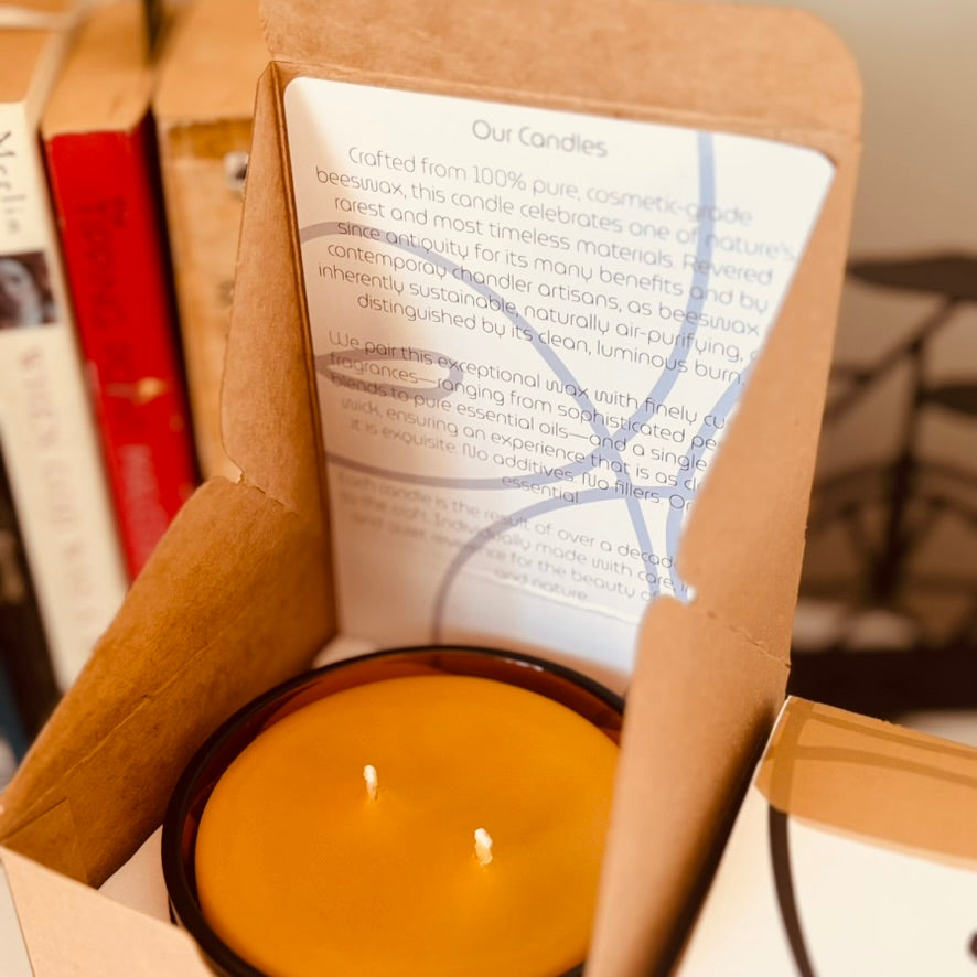 Candle in a cardboard box with a label inside, set against a blurred bookshelf background.
